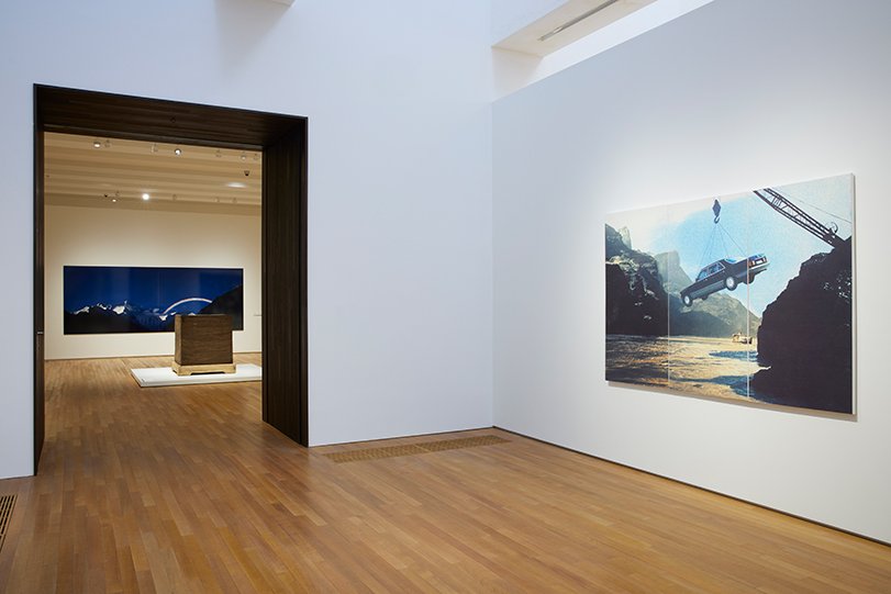 A canvas print is hung on the white wall of a gallery and features a car hoisted over a river by a crane. Towards the left of the work is a doorway framed in black leading to the next gallery. There, a cube-like sculpture is situated on the ground before a wall display of a photograph of a rainbow.
